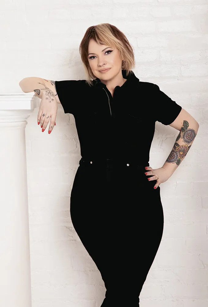 Person with tattoos and short hair leans against a white pillar, wearing a black outfit, in front of a white brick wall. - The Wayward Hairdresser | Brooklyn, NY